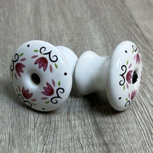 May include: Two white ceramic cabinet knobs with a floral design. The knobs feature a central hole and are decorated with pink flowers, green leaves, and black accents. The knobs are approximately 1.5 inches in diameter.