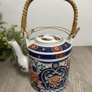 May include: A ceramic teapot with a bamboo handle. The teapot features a white base with blue and orange floral patterns. The handle is made of woven bamboo. The spout and lid are also white with floral accents. The teapot is designed for serving tea.