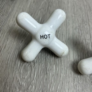 Peut inclure: Poignée de robinet en céramique blanche en forme de croix. Le mot "HOT" est imprimé en noir sur la poignée. La poignée est conçue pour un robinet de salle de bain ou de cuisine.