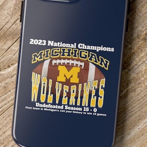 May include: A blue phone case with a white and yellow Michigan Wolverines logo and the text "2023 National Champions Undefeated Season 15-0 First team in Michigan's 144 year history to win 15 games."