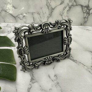 May include: Ornate silver-toned picture frame with a rectangular opening. The frame features detailed scrollwork and floral accents. The frame is designed to hold a photograph or artwork. The frame is displayed on a marble surface.