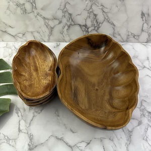 Pode incluir: Um conjunto de tigelas de madeira com acabamento natural de grão de madeira. As tigelas têm um design de borda ondulada. Uma tigela grande e uma pilha de tigelas menores são visíveis. As tigelas estão dispostas sobre uma superfície com padrão de mármore.