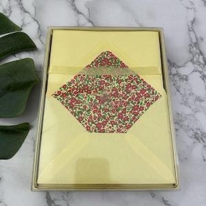 May include: A stationery set in a clear box. The set includes pale yellow notecards and envelopes. The envelope lining features a floral pattern with red flowers and green leaves on a cream background. A yellow ribbon is tied around the set.