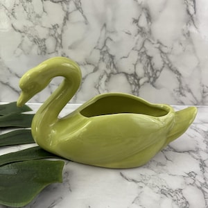 May include: A glossy, light green ceramic swan-shaped planter or decorative bowl. The swan features a long, curved neck, a smooth body with an open top cavity, and a pointed tail. The finish is uniform and reflective, highlighting the curves.