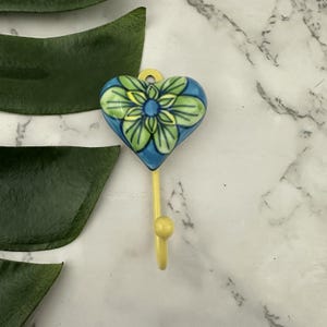 May include: A heart-shaped ceramic wall hook with a floral design. The hook features a blue background with a green and yellow flower design. The hook itself is yellow and has a small ball at the end. The hook is approximately 7.6 cm long.