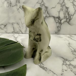 May include: A ceramic cat figurine in a seated position. The cat is primarily off-white with dark green and black marbling. A floral design is visible on the cat's chest. The figurine is on a white marble surface.