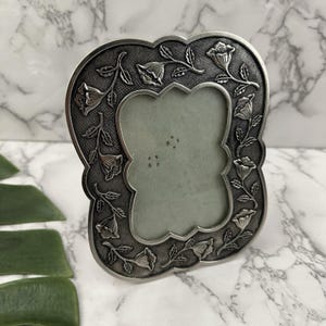 May include: A silver-toned picture frame with a floral design. The frame has a scalloped edge and is decorated with detailed embossed flowers and leaves. The frame is designed to hold a small photograph or artwork.