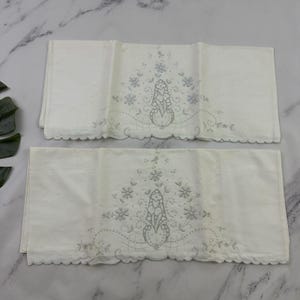Puede incluir: Dos piezas de tela rectangulares de color blanco con motivos florales bordados en gris claro en el centro. El bordado incluye delicados patrones de flores y hojas con bordes festoneados en la parte inferior de ambas piezas.