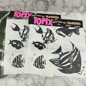 May include: Two packages of Topix easy iron-on transfers featuring various black and white fish designs. The fish include striped and patterned varieties, with the Topix logo in pink and black.