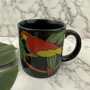 May include: Black ceramic mug featuring a colorful parrot design. The parrot has red, yellow, and green details, perched on a branch with green leaves. The mug has a curved handle and a glossy finish.