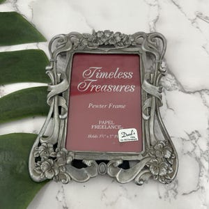 May include: A silver pewter picture frame with an ornate design featuring floral accents. The frame has a red mat and the text "Timeless Treasures Pewter Frame" is printed on the mat. The frame is labeled "Papel Freelance" and holds a 3 1/2 x 5 inch picture.