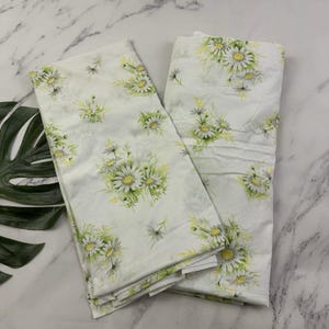 Puede incluir: Dos sábanas blancas con un estampado floral de margaritas. Las sábanas presentan racimos de margaritas blancas con centros amarillos y hojas verdes. El fondo es blanco y las sábanas están dobladas.