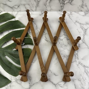May include: An expandable wooden wall rack featuring a brown finish and ten pegs arranged in a diamond-shaped lattice pattern. The rack is designed with a folding accordion mechanism that allows for adjustable width and multiple hanging points.