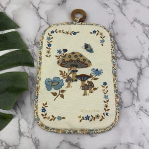 Könnte beinhalten: Ein rechteckiger Topflappen aus Stoff mit cremefarbenem Hintergrund, einem braun-blauen Pilz- und Blumenmuster, einem kleinen blauen Schmetterling und einem hölzernen Aufhängering. Der Text David Davir ist unten rechts auf den Stoff gedruckt.