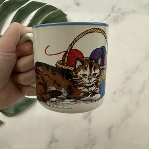Puede incluir: Una taza de cerámica blanca con un borde azul presenta un gato atigrado marrón con ojos verdes. El gato está rodeado de una cesta de ovillos de lana de colores.