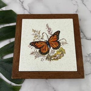 May include: Square wooden framed tile featuring a monarch butterfly with orange and black wings. The tile has a cream-colored background with a floral and landscape design. The frame is a dark brown color.