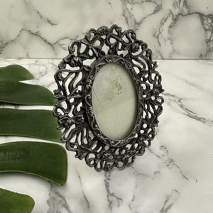 May include: An oval, antique-style picture frame with an ornate, scrolling design. The frame is made of a dark, possibly pewter-toned metal. The frame's interior holds a light-colored backing, ready for a photograph. The frame is displayed on a marble surface.