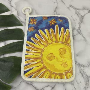 May include: A rectangular fabric pot holder featuring a yellow sun with a human face surrounded by golden rays against a dark blue starry night sky. The item is bordered by white piping and includes a small white fabric loop at the top corner.