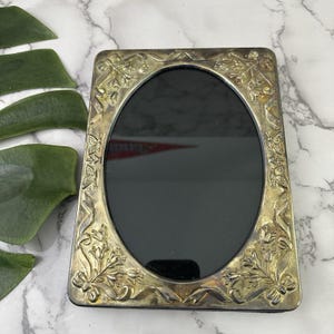 May include: A rectangular silver-toned picture frame with an oval opening. The frame is decorated with floral and ribbon designs. The background is a white and gray marble surface.
