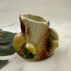 May include: A ceramic planter shaped like a tree stump with three yellow mushrooms attached. The planter is a light brown color with a cream-colored interior.