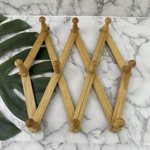 May include: A light-colored wooden accordion-style coat rack with multiple pegs. The rack is made of thin, rectangular wooden slats joined in a zig-zag pattern. The pegs are round and extend outward. The rack is on a white and gray marble-patterned surface.