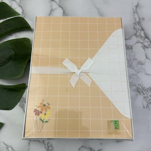 May include: A rectangular gift box with a peach-colored grid pattern and a white diagonal design. A white ribbon is tied in a bow around the box. A small floral design is on the lower left corner.