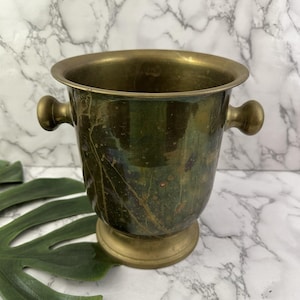 May include: A brass wine cooler featuring a wide, flared rim, a tapered body with a weathered, dark patina, and two rounded side handles. The vessel stands on a circular pedestal base, showcasing a metallic finish with visible surface oxidation.