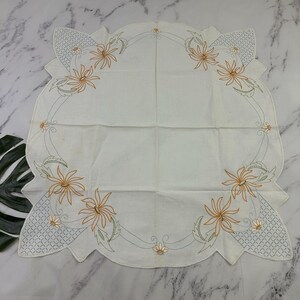 Puede incluir: Un tapete cuadrado de lino blanquecino con borde festoneado. El tapete presenta un diseño floral bordado en naranja, verde y gris. El diseño incluye flores y hojas estilizadas, con un patrón geométrico en las esquinas.
