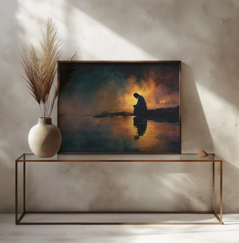 May include: A framed art print showing a person kneeling by water, set against a dramatic sky in orange, blue, and yellow hues. The artwork is displayed on a glass-topped table with a bronze metal frame, next to a vase with dried ornamental grass.