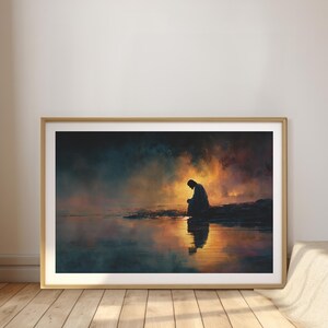 May include: Framed art print showing a silhouetted figure kneeling by water, with a fiery, colourful sky reflected in the water. The artwork is set in a gold-coloured frame and rests on a wooden floor.