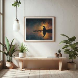May include: A framed art print showing a silhouette of a person by water, set against a colourful sunset. The artwork hangs above a wooden bench with potted plants, creating a calm and reflective atmosphere.
