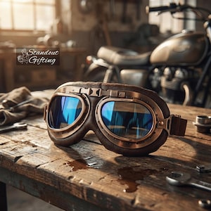 May include: Vintage-style brown leather goggles with copper-colored metal frames and blue-tinted lenses. The goggles are resting on a wooden surface with tools and a motorcycle in the background. The text "Standout Gifting" is visible.