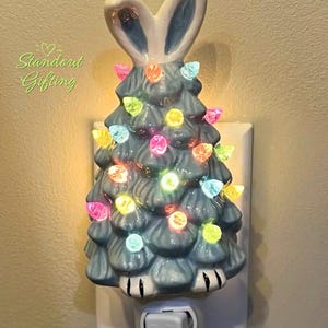 May include: A ceramic night light shaped like a tree with bunny ears, painted in shades of blue and white. The tree is adorned with colorful lights. The night light is plugged into a wall outlet and has an on/off switch. The text "Standout Gifting" is visible.