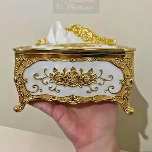 May include: An ornate, rectangular tissue box with a white and gold design. The box features a white front panel with gold rose detailing and a gold filigree border. The top and feet are also gold. The text "Standout Gifting" is in the upper right corner.