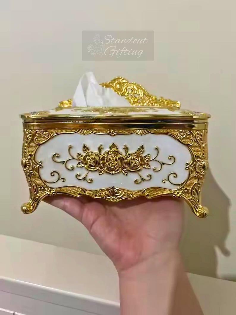 May include: An ornate, rectangular tissue box with a white and gold design. The box features a white front panel with gold rose detailing and a gold filigree border. The top and feet are also gold. The text "Standout Gifting" is in the upper right corner.