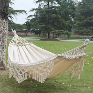 May include: A cream-coloured hammock with a macrame fringe hangs between two trees in a grassy area. The hammock is made of a soft, woven fabric, designed for outdoor relaxation. The background features trees and a cloudy sky.