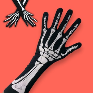 Può includere: Guanti neri con un design di ossa di scheletro bianche. I guanti si estendono sul braccio e presentano una stampa dettagliata di ossa su mani e avambracci. Lo sfondo è di colore corallo.