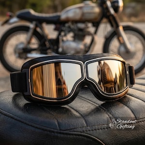May include: Black and silver motorcycle goggles with reflective lenses. The goggles have a black leather frame and a silver trim. The background features a vintage motorcycle.