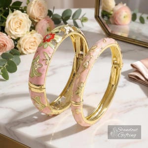May include: Two gold-toned hinged bangles with pink enamel floral designs. One bangle features red and green floral accents. The other bangle has a simpler design. The bangles are displayed on a marble surface.