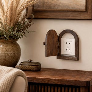 May include: A wooden electrical outlet cover with an arched frame and a small door. The door is open, revealing a white outlet. A decorative vase with dried plants and a small, ornate box are on a wooden surface.