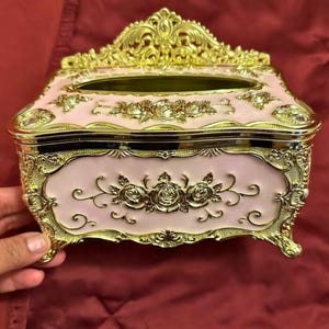 May include: A decorative tissue box in a rectangular shape, featuring a light pink body with gold-colored ornate detailing. The box has a floral design and is elevated on small feet. The top has an opening for tissue dispensing.