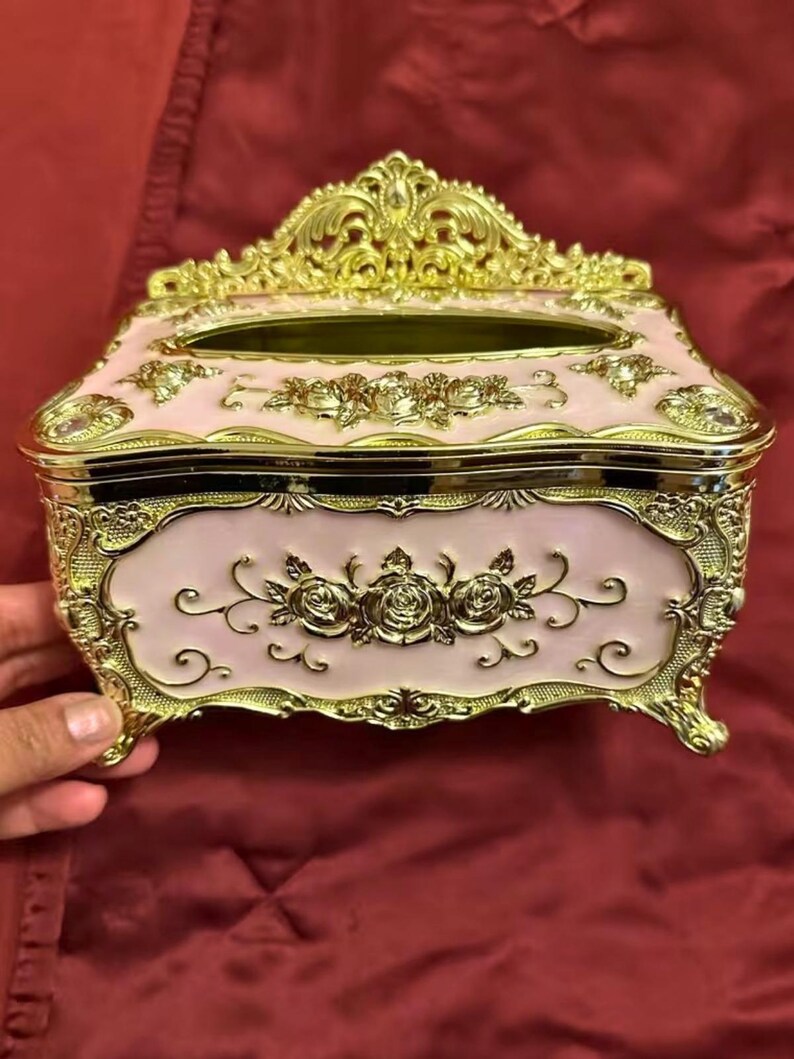 May include: A decorative tissue box in a rectangular shape, featuring a light pink body with gold-colored ornate detailing. The box has a floral design and is elevated on small feet. The top has an opening for tissue dispensing.