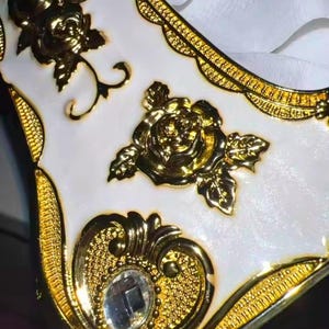 May include: Close-up of a white and gold decorative box. The box features gold floral embellishments, including roses, and a decorative border. A clear, square-cut gem is set into the gold filigree design.