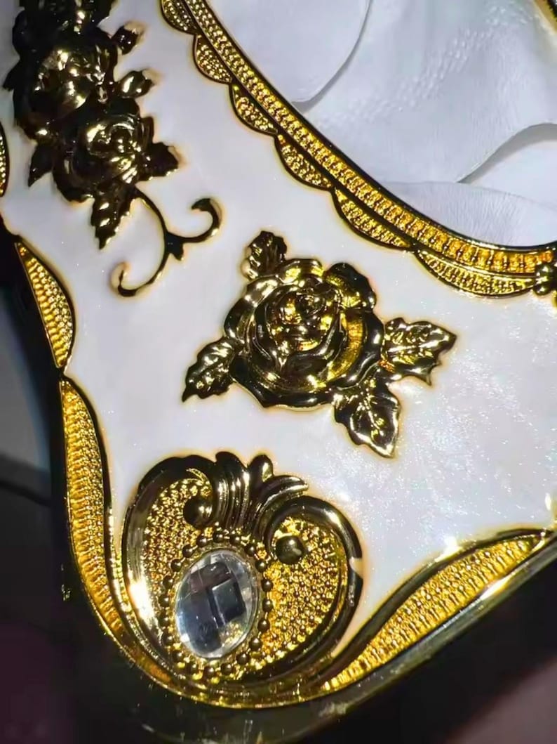 May include: Close-up of a white and gold decorative box. The box features gold floral embellishments, including roses, and a decorative border. A clear, square-cut gem is set into the gold filigree design.