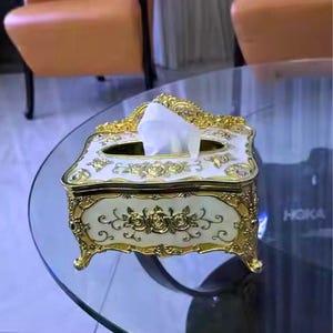 May include: An ornate tissue box with a cream-colored base and gold accents. The box features floral designs and is placed on a glass table. The design is reminiscent of vintage decor.