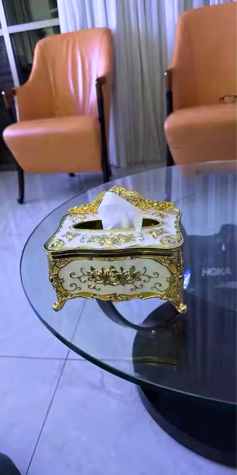 May include: An ornate tissue box with a cream-colored base and gold accents. The box features floral designs and is placed on a glass table. The design is reminiscent of vintage decor.