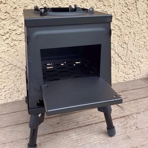 Puede incluir: Una estufa de leña portátil negra y rectangular con una puerta con bisagras y patas ajustables. La estufa tiene una chimenea y una rejilla en el interior. La estufa está diseñada para uso en exteriores y acampada.