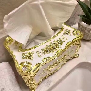May include: An ornate tissue box in white and gold. The box features a floral design with gold trim and accents. White tissues are visible, pulled from the opening. The box is on a marble surface, with a small plant in the background.