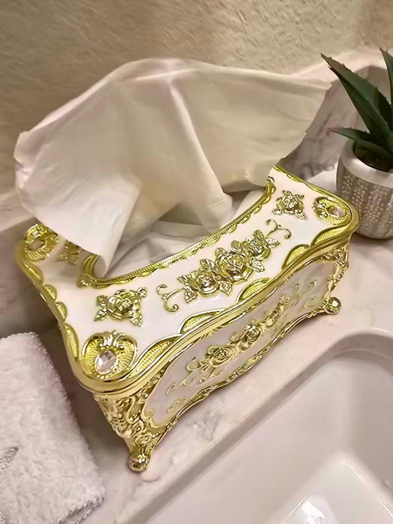 May include: An ornate tissue box in white and gold. The box features a floral design with gold trim and accents. White tissues are visible, pulled from the opening. The box is on a marble surface, with a small plant in the background.