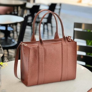May include: A brown leather tote bag with two top handles and a detachable shoulder strap. The bag has a rectangular shape with a zipper closure and vertical seams on the front and back. The bag is sitting on a white surface.